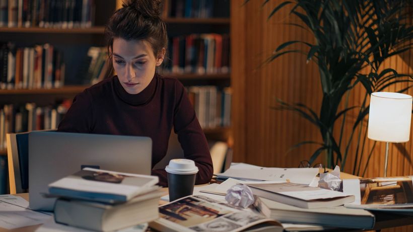 Woman working late at a desk filled with books, laptop, and coffee in a cozy library setting