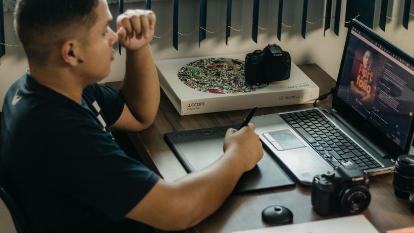 A photographer working at a creative setup with a laptop and Wacom tablet, focused on editing photos indoors