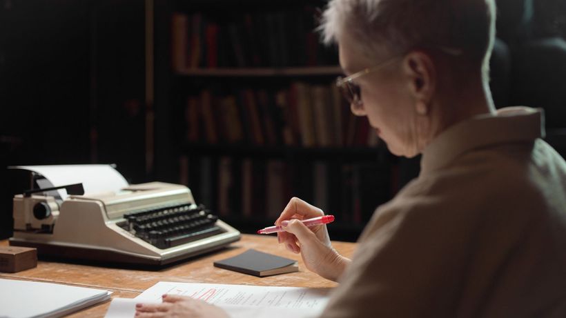 Elderly woman reviewing a manuscript with a typewriter at a wooden desk