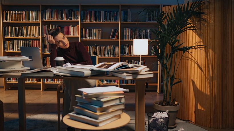 A young woman sits at a cluttered desk in a library, surrounded by books and a laptop