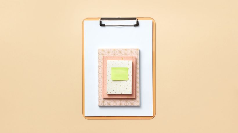 Stylish flat lay of a clipboard with pastel notebooks and a green sticky note on a beige background
