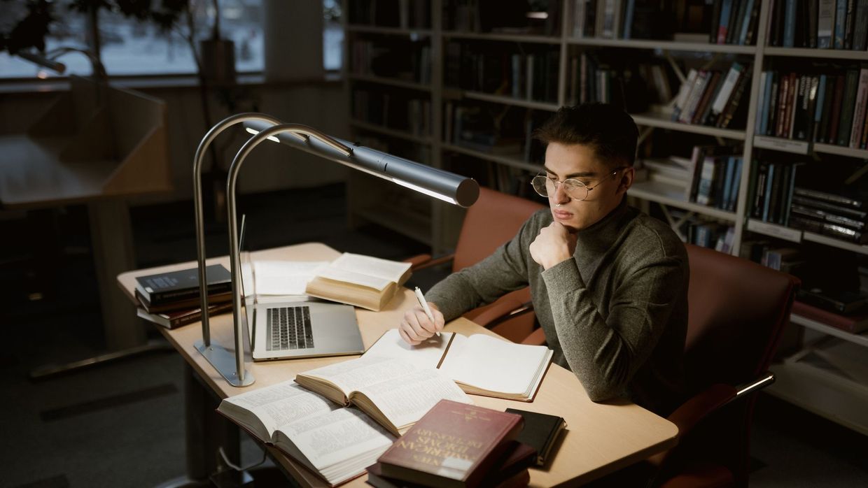 Free SEO courses: a young adult studying at a library desk with books and laptop under warm lighting