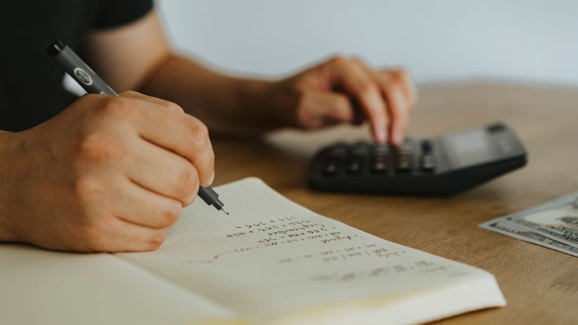 Close-up of hands writing calculations in a notebook with a calculator, focused on budgeting or financial work