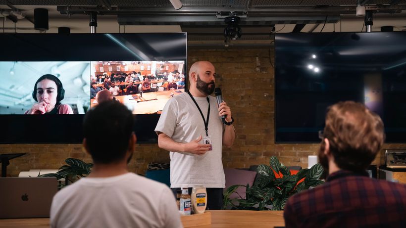 Speaker presenting in a modern office with audience and virtual call on screens