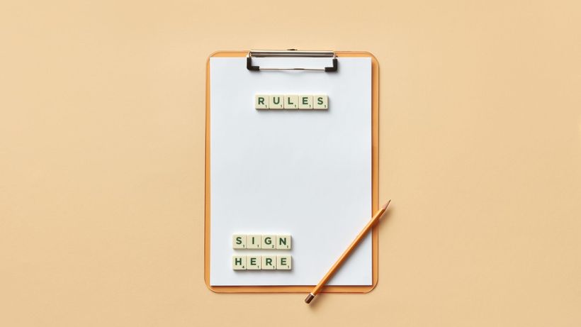Flat lay of clipboard with letter tiles spelling 'rules' and 'sign here', accompanied by pencil on beige background