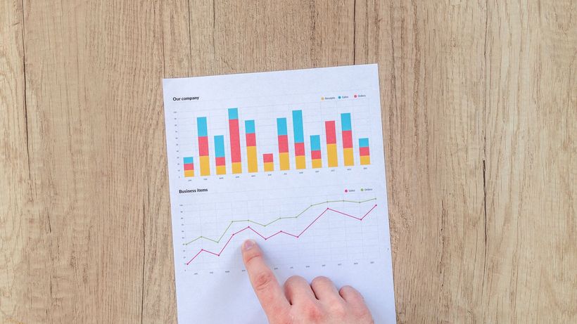 A hand points to colorful business charts and graphs on a paper sheet on a wooden desk
