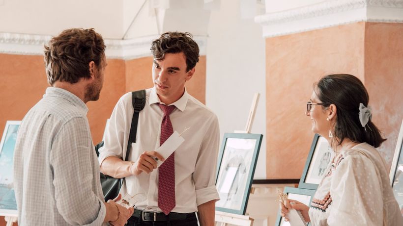 Three adults engaged in conversation at an art gallery exhibition with paintings on display