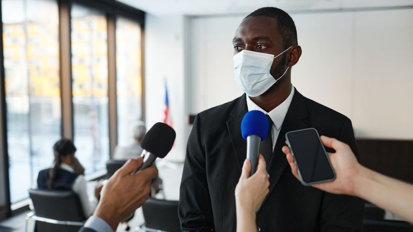 African American businessman in formal attire speaks to media while wearing a face mask in a conference room setting