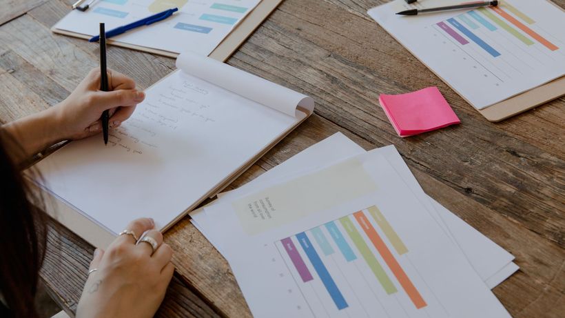 Hands writing on paper with colorful charts on wooden table, organized workspace