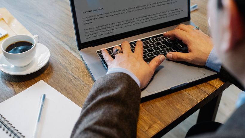 Crop faceless male entrepreneur in formal wear sitting at table with cup of coffee and notebook while working on project