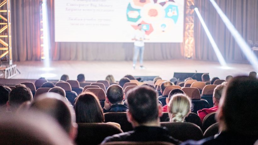 Audience attentively watching a presentation in a conference setting, with stage lights and speaker visible