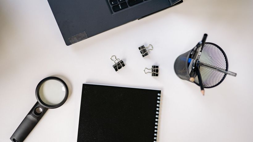 Flat lay of a workspace featuring a laptop, black notebook, magnifying glass, and office supplies on a white surface