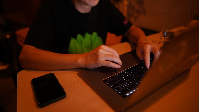 An adult man working at a desk on a laptop with a smartphone nearby, in a dimly lit home office