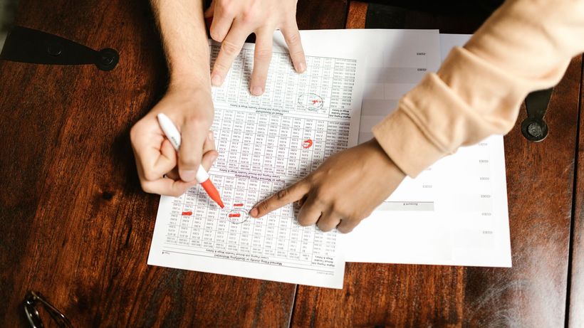 Two hands reviewing and marking financial documents with a red pen on a wooden table, top view