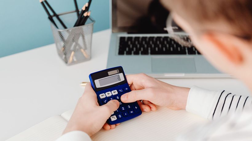 Close-up of hands using a blue calculator at a desk with a laptop and stationery