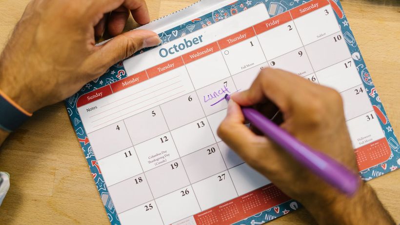 Close-up of a person writing a lunch reminder on an October calendar with a purple pen