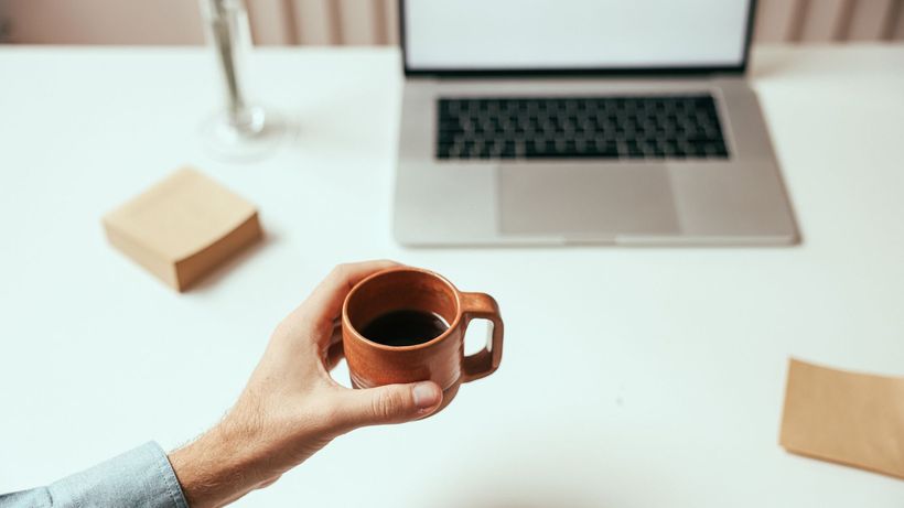 A person holds a brown coffee cup near a laptop on a clean white desk, perfect for remote work themes