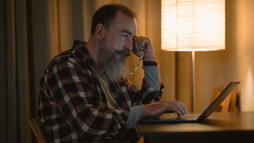 Elderly man with a beard working late on laptop under warm lamp light indoors