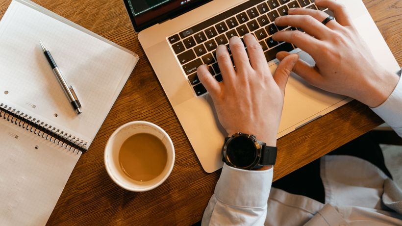 Hands typing on a laptop next to a cup of coffee and a notebook on a wooden table