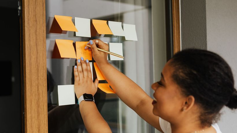 A woman writes notes on a window using colorful sticky notes, emphasizing planning and creativity
