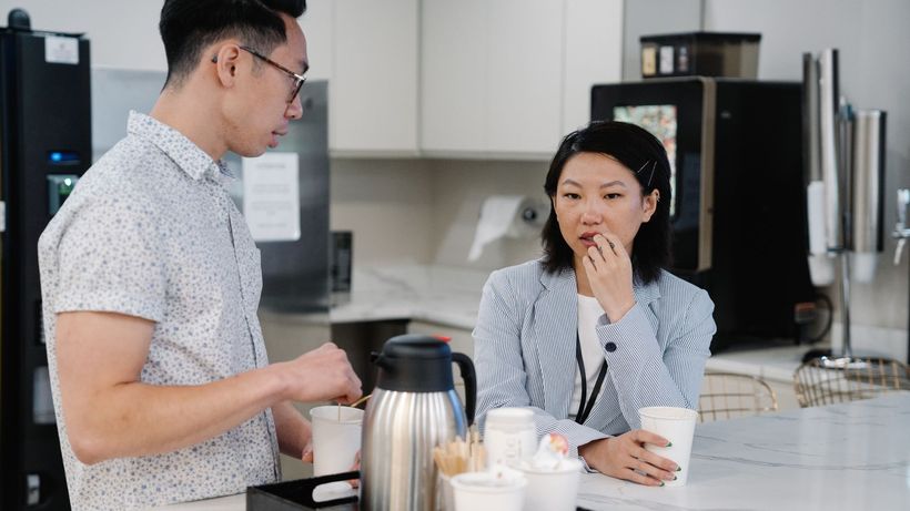 Two colleagues discussing over coffee in a modern office break room setting