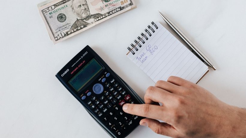Top view of crop unrecognizable economist calculating on calculator on table with notepad and pen near pile of dollar