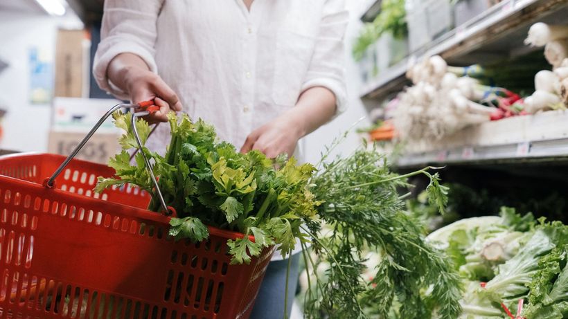 A person holding a grocery basket filled with fresh greens like celery and parsley in a store