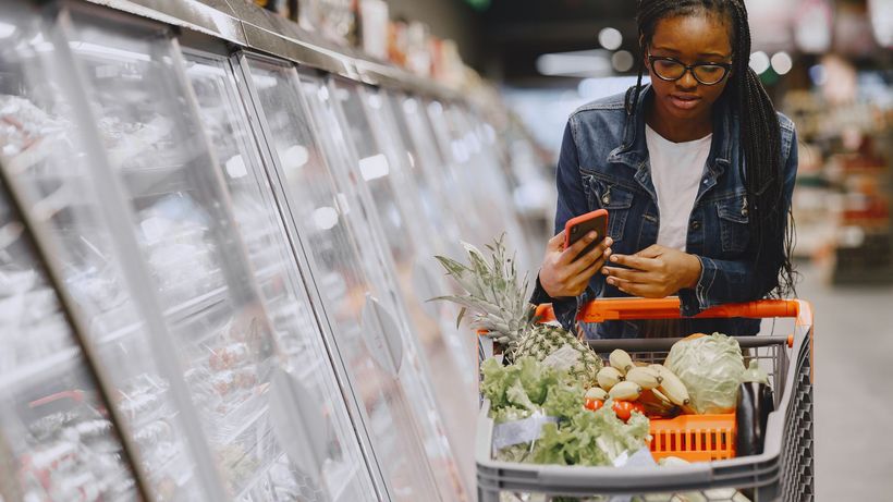 Woman in a grocery store using smartphone while pushing a shopping cart filled with groceries