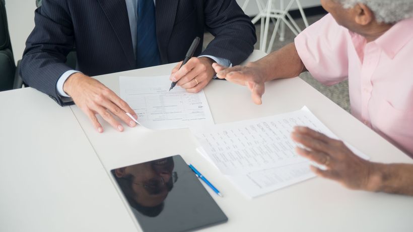 A business meeting between an elderly client and a consultant discussing documents at an office table