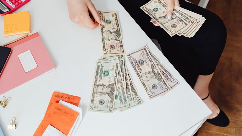 A person counting US dollar bills at a white desk with office supplies nearby