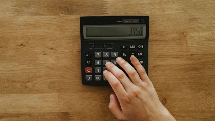 A person's hand pressing keys on a calculator displaying 3750 on a wooden table, top view
