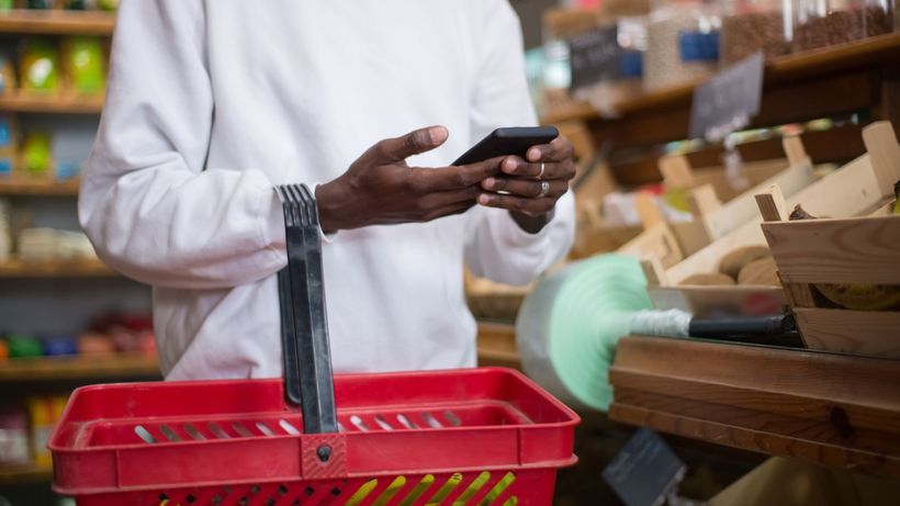 A man shops using a smartphone at a grocery store, holding a red basket filled with groceries