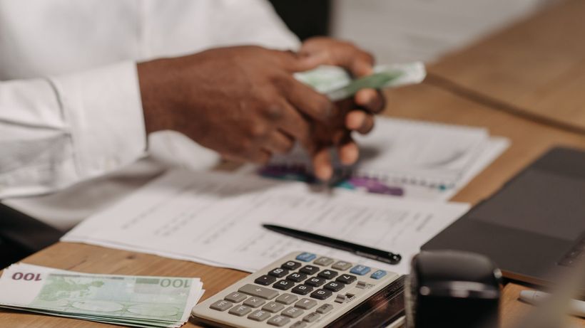 Hands counting euro bills on a wooden desk with calculator, financial documents, and laptop nearby