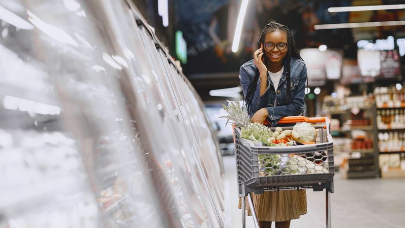 Smiling woman multitasking by shopping with a cart in a supermarket aisle while talking on the phone