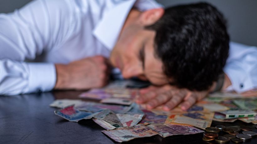 A man in a white shirt rests his head on a table covered with Argentine pesos and coins