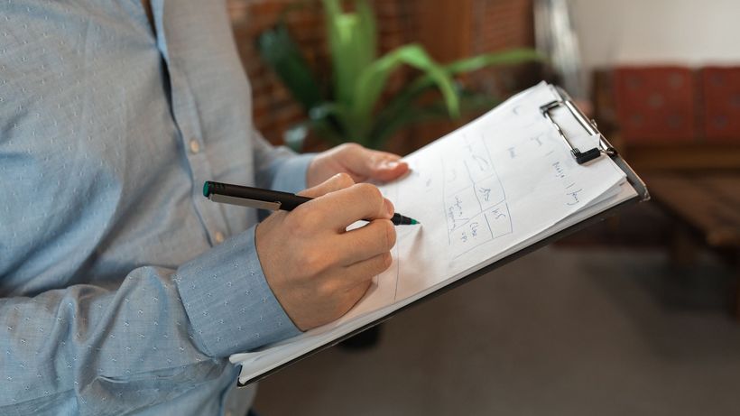 A person in blue shirt writes with a pen on a clipboard indoors, showcasing focus and productivity