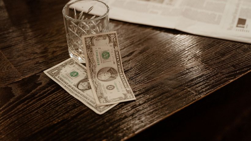 Close-up view of dollar bills with a glass on a wooden table, beside a newspaper