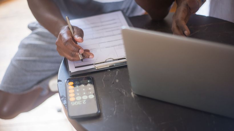 A person working on a laptop with a smartphone and clipboard on a round table