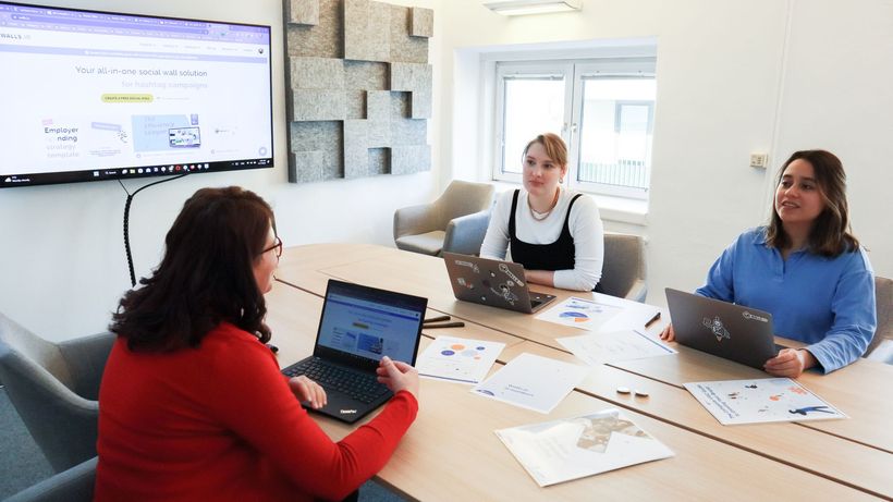 Three women collaborating at a conference table in a modern office with laptops and presentation screen